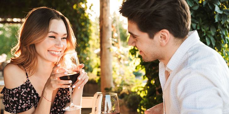 Picture of young loving couple sitting in cafe by dating outdors in park holding glasses of wine drinking talking with each other.