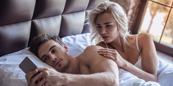 Beautiful young couple is lying in bed guy is using a smart phone girl is looking with reproach