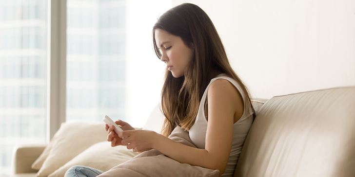 Sad teen girl using cellphone while sitting on sofa at home, writing message, typing contact, reading posts in social network. Upset young woman procrastinates or bored, playing with mobile phone